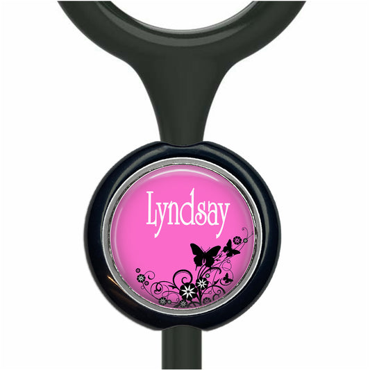 A pink circular charm with the name "Lyndsay" written on it, surrounded by black floral designs.