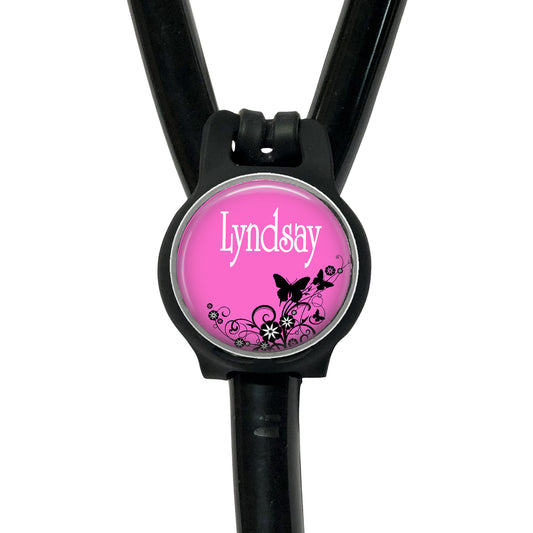 A pink-colored circular badge with the name "Lyndsay" printed on it, attached to a black stethoscope.