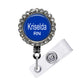 A blue badge with the name "Krisela RN" printed on it, surrounded by a decorative metal frame with rhinestones.