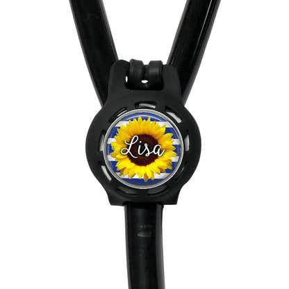 A black stethoscope with a personalized nameplate attached to it. The nameplate features a sunflower design and the name "Lisa".